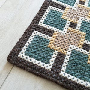 Close-up of a brown, green, and beige woven rug with a geometric pattern. The rug has a white border around each geometric shape.