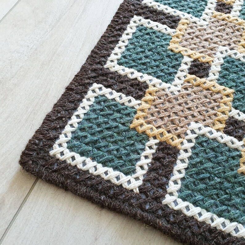 Close-up of a brown, green, and beige woven rug with a geometric pattern. The rug has a white border around each geometric shape.