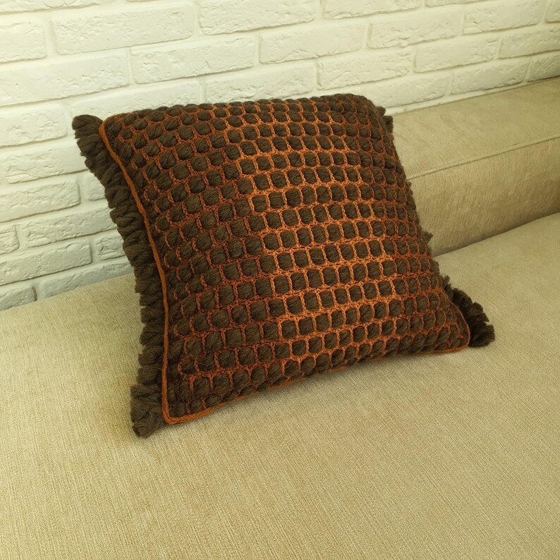 Puede incluir: Un coj&iacute;n decorativo cuadrado con un patr&oacute;n texturizado en forma de rejilla en tonos marr&oacute;n y naranja. El coj&iacute;n tiene un borde con flecos y est&aacute; colocado sobre un sof&aacute; beige, ofreciendo un acento acogedor y elegante para la decoraci&oacute;n del hogar.