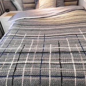 A gray knitted throw blanket with a checkered pattern in black, white, and blue. The blanket is made of a soft, textured yarn and has a relaxed, casual look.
