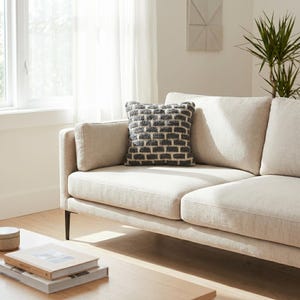 May include: A neutral-toned sofa with a cushion displaying a grey and white brick design. The sofa has a contemporary style with dark legs. The room is well-lit, with a window and a plant visible.