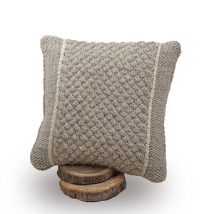 May include: A square, light grey knitted cushion with a textured weave pattern. The cushion has a lighter-coloured border on each side and is displayed on a small wooden base. The cushion is approximately 35 cm square.