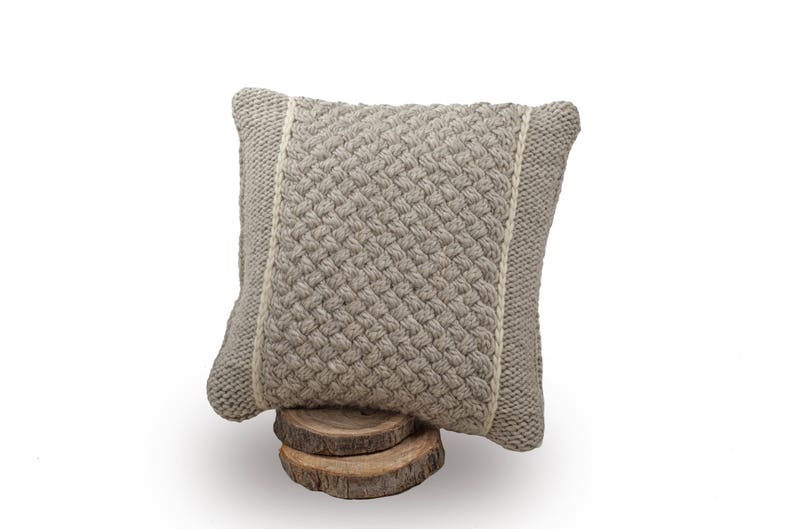 May include: A square, light grey knitted cushion with a textured weave pattern. The cushion has a lighter-coloured border on each side and is displayed on a small wooden base. The cushion is approximately 35 cm square.
