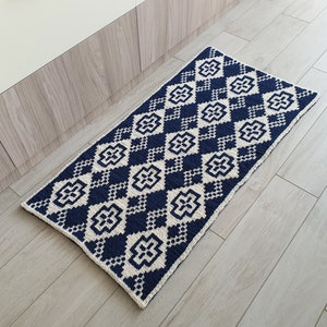 May include: A navy blue and white geometric patterned rug with a diamond pattern. The rug is made of a soft, woven material and has a fringed edge.