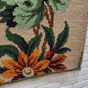 A close-up of a beige tapestry with a floral design. The tapestry features green leaves, brown stems, and orange and brown flowers. The tapestry has a fringe border.