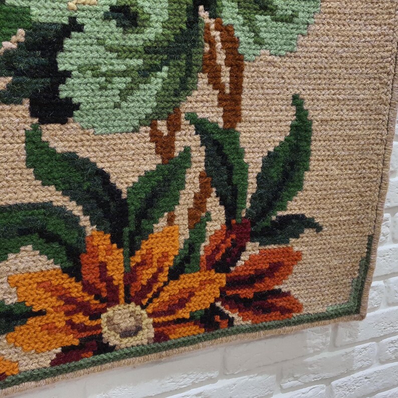 A close-up of a beige tapestry with a floral design. The tapestry features green leaves, brown stems, and orange and brown flowers. The tapestry has a fringe border.