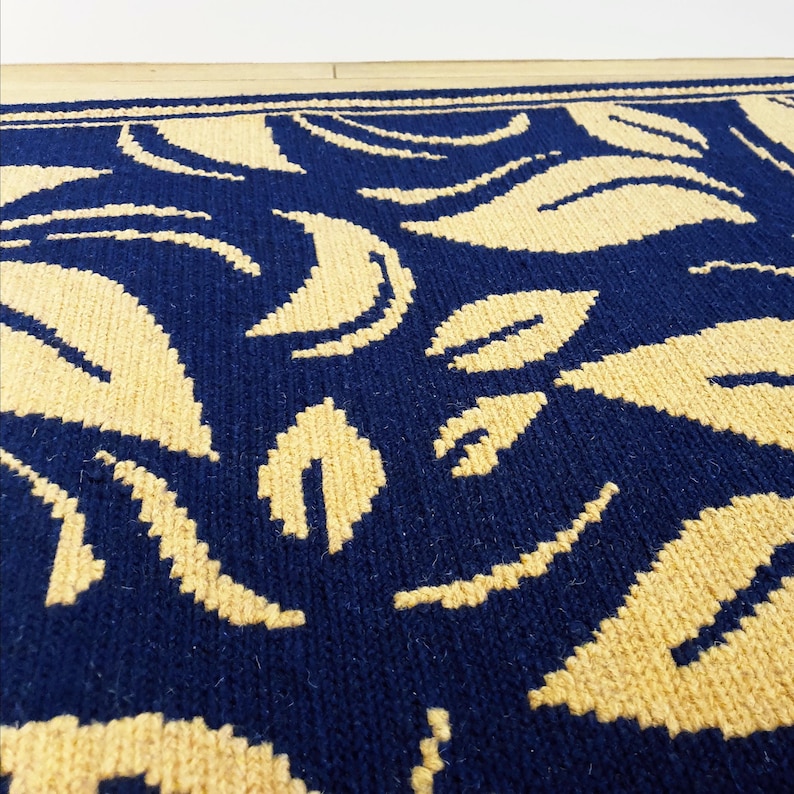 A blue and beige patterned rug with a woven texture. The rug features a repeating pattern of stylized leaves.