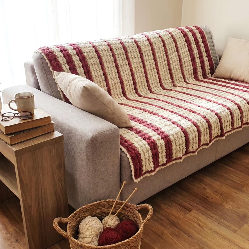 A white and pink striped crocheted throw blanket with fringe on a gray couch. The blanket has a textured, bumpy design.