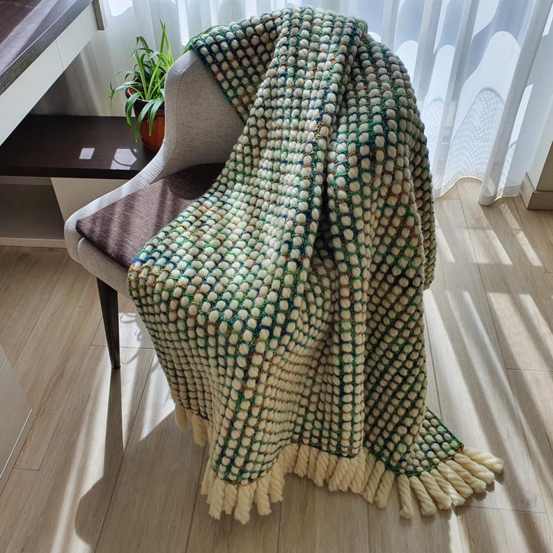 A textured, green and cream-colored throw blanket with a dotted pattern is draped over a chair. The blanket has a fringe trim at the bottom. The blanket is made of a soft material and is perfect for adding warmth and style to any room.