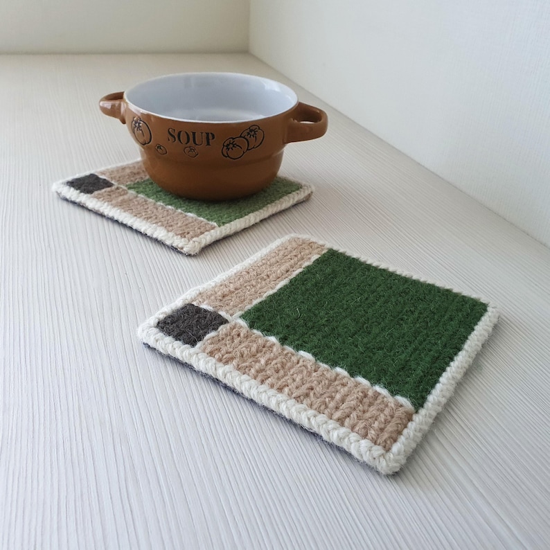 May include: Two handmade crochet coasters with a geometric design. Each coaster features a dark green square, a tan rectangle, and a dark gray square, bordered by a white trim. A brown soup bowl sits on one coaster.
