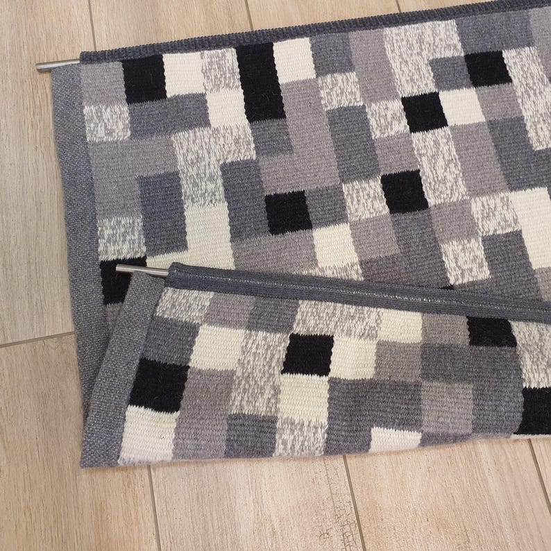 K&ouml;nnte beinhalten: Ein grauer und wei&szlig;er karierter Teppich mit schwarzen Quadraten. Der Teppich hat einen grauen Rand und h&auml;ngt an einer Metallstange.
