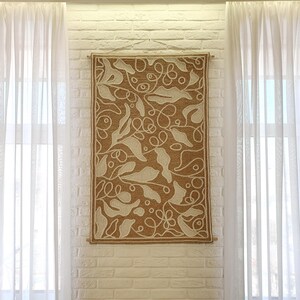 Decorative wall panel with wool stitching, textured surface and abstract leaf motif on a wall in a modern living room between two windows.