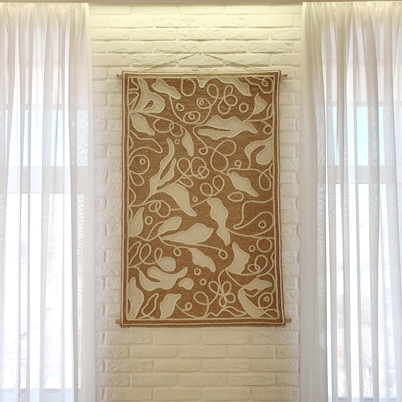 Decorative wall panel with wool stitching, textured surface and abstract leaf motif on a wall in a modern living room between two windows.
