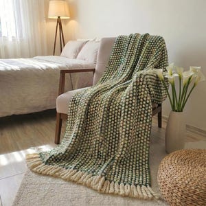 Puede incluir: Una manta texturizada verde y crema con flecos est&aacute; sobre una silla. La manta tiene un patr&oacute;n de puntos y est&aacute; hecha de un material suave. La silla es de color neutro y est&aacute; en un dormitorio.