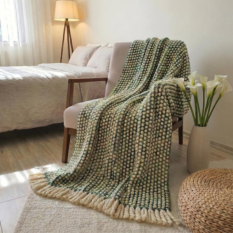 Puede incluir: Una manta texturizada verde y crema con flecos est&aacute; sobre una silla. La manta tiene un patr&oacute;n de puntos y est&aacute; hecha de un material suave. La silla es de color neutro y est&aacute; en un dormitorio.