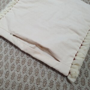 May include: Cream-colored pillow with a textured, corduroy-like surface. The pillow has a hidden zipper and is trimmed with a thick, fluffy, white fringe. The pillow is resting on a patterned, neutral-toned surface.
