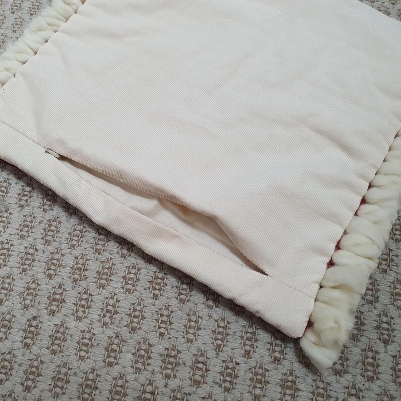 May include: Cream-colored pillow with a textured, corduroy-like surface. The pillow has a hidden zipper and is trimmed with a thick, fluffy, white fringe. The pillow is resting on a patterned, neutral-toned surface.