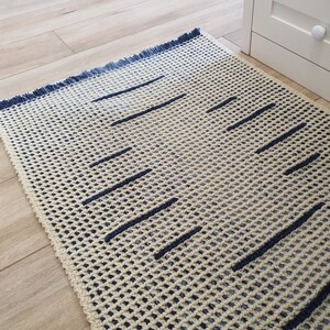 May include: A cream-colored rug with a blue fringe and a geometric pattern of dark blue lines. The rug is made of a woven material and has a textured surface.