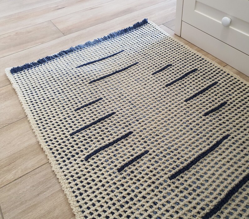 May include: A cream-colored rug with a blue fringe and a geometric pattern of dark blue lines. The rug is made of a woven material and has a textured surface.