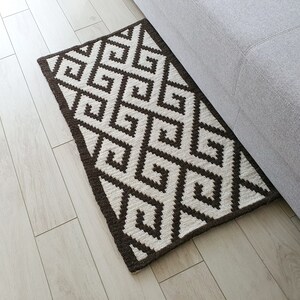 May include: A brown and white geometric patterned rug with a diamond design. The rug is made of woven fibers and has a fringed edge.