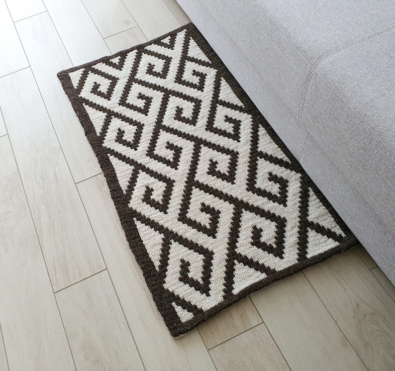May include: A brown and white geometric patterned rug with a diamond design. The rug is made of woven fibers and has a fringed edge.