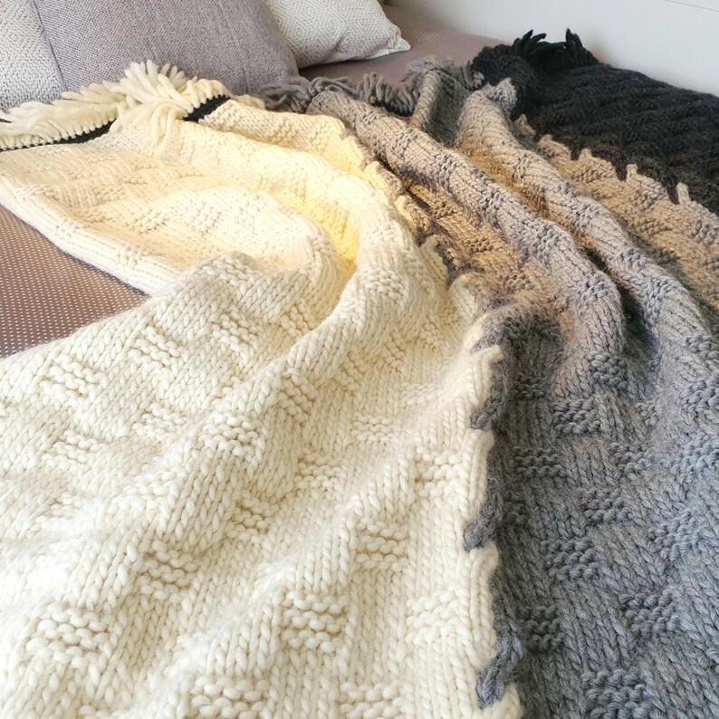 A white and gray knitted throw blanket with a fringe border. The blanket is made of chunky yarn and has a textured pattern.