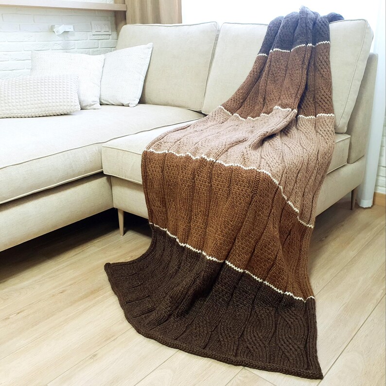 A brown, beige, and white striped knitted throw blanket draped over a beige couch. The blanket has a cable knit pattern.