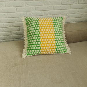 May include: A square decorative pillow with a green and white polka dot pattern on the sides and a yellow center. The pillow has a fringe trim and is placed on a beige surface.