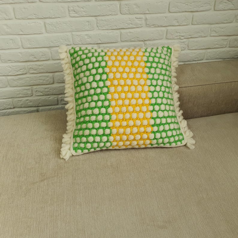 May include: A square decorative pillow with a green and white polka dot pattern on the sides and a yellow center. The pillow has a fringe trim and is placed on a beige surface.