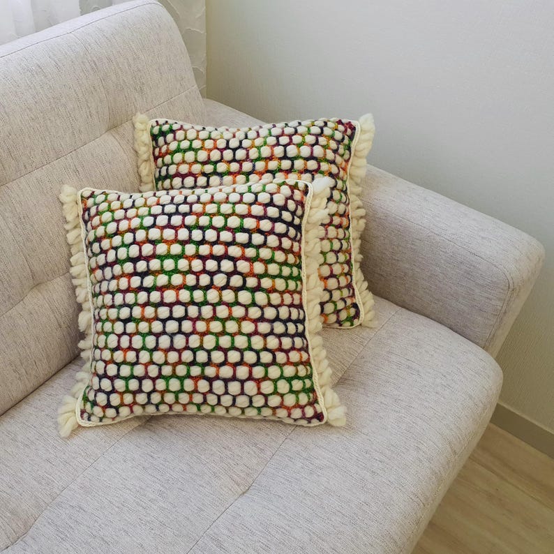 K&ouml;nnte beinhalten: Zwei quadratische Zierkissen mit einem strukturierten, mehrfarbigen Muster aus wei&szlig;en, gr&uuml;nen, roten und lila Punkten. Die Kissen haben eine cremefarbene Fransenborte und liegen auf einem hellgrauen Sofa.