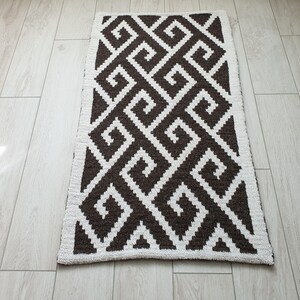 May include: A brown and white geometric patterned rug with a white border. The rug has a repeating pattern of squares and swirls.