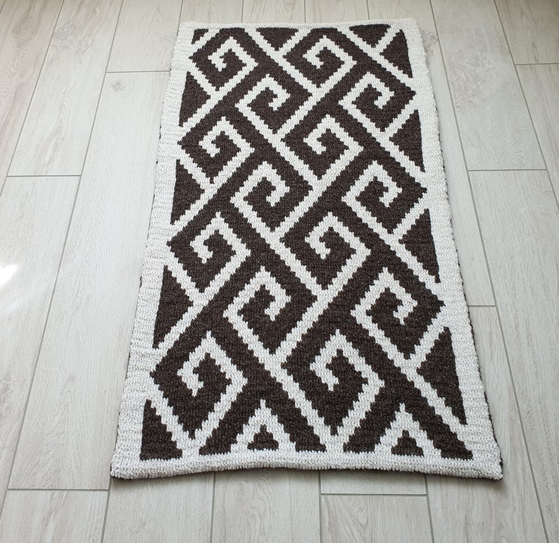 May include: A brown and white geometric patterned rug with a white border. The rug has a repeating pattern of squares and swirls.