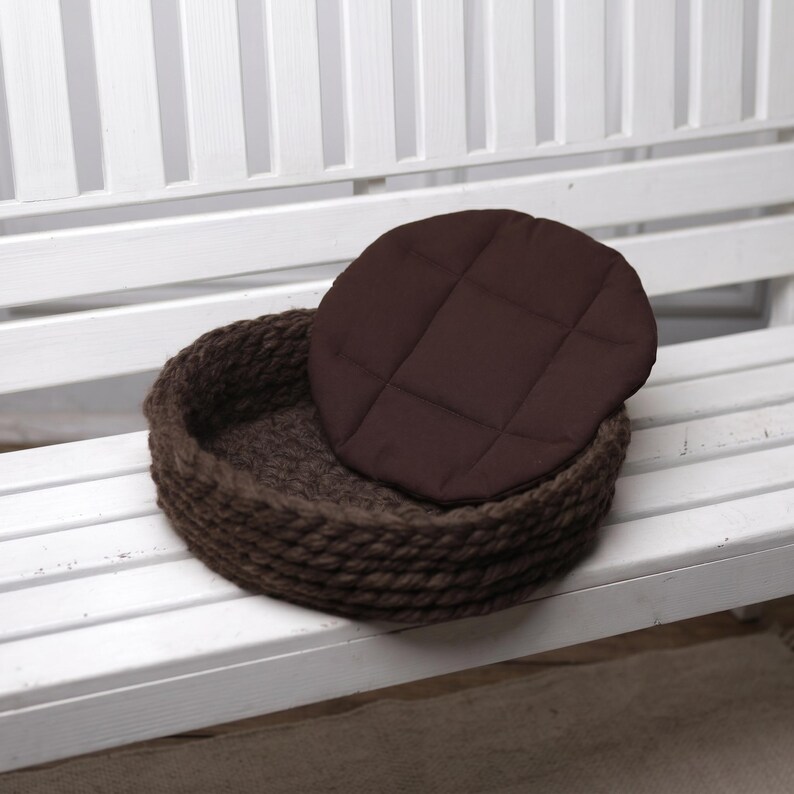 A round dark brown hand-knitted wool cat bedding with a chunky yarn texture, shown with its removable quilted fabric cushion leaning against the side, displayed on a white wooden bench next to a white radiator.