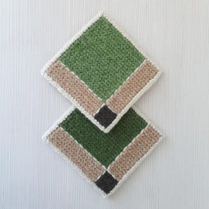May include: Two handmade crochet coasters. Each coaster is square with a green center, a tan border, and a dark gray square in the corner. The edges are finished with a white border. The coasters are displayed on a white surface.