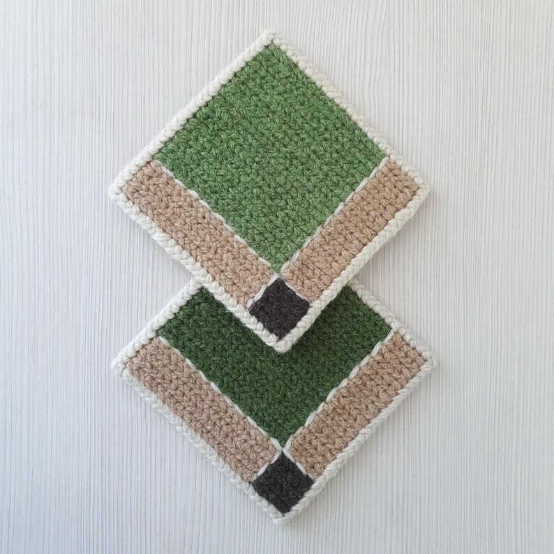 May include: Two handmade crochet coasters. Each coaster is square with a green center, a tan border, and a dark gray square in the corner. The edges are finished with a white border. The coasters are displayed on a white surface.
