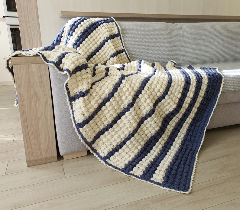 May include: A white and blue striped crocheted throw blanket with a textured bobble stitch. The blanket is draped over a couch.