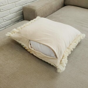 May include: A square, cream-colored decorative pillow with a zipper, revealing a white pillow insert. The pillow has a textured, off-white fringe trim around the edges. The pillow is resting on a beige sofa.