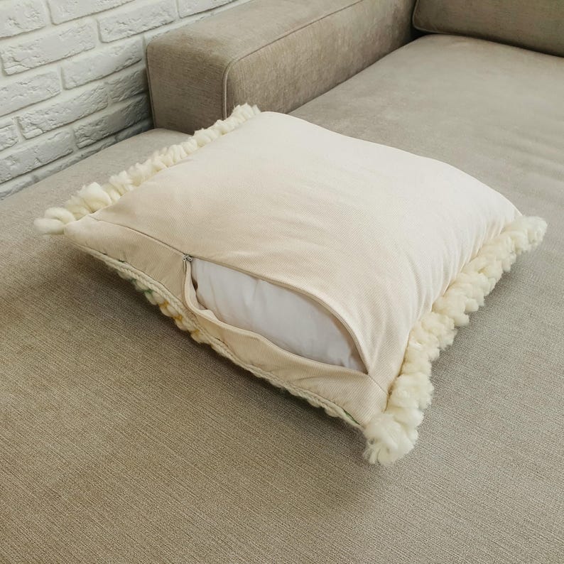 May include: A square, cream-colored decorative pillow with a zipper, revealing a white pillow insert. The pillow has a textured, off-white fringe trim around the edges. The pillow is resting on a beige sofa.