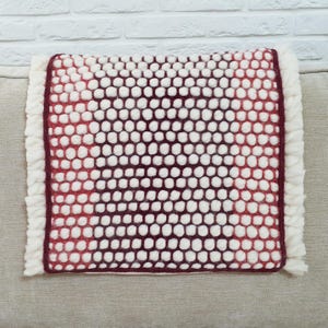 May include: A rectangular pillow with a textured surface of white and burgundy felted wool balls. The pillow has a burgundy border and a white fringe on the edges. The pillow is on a neutral-colored surface.