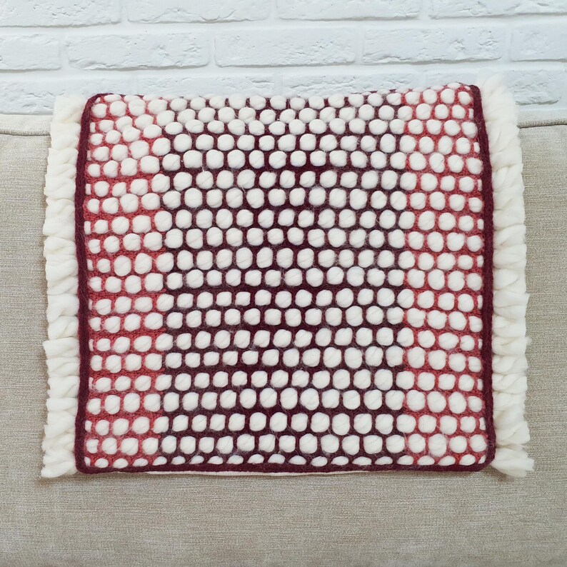 May include: A rectangular pillow with a textured surface of white and burgundy felted wool balls. The pillow has a burgundy border and a white fringe on the edges. The pillow is on a neutral-colored surface.