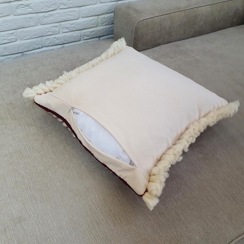May include: Cream-colored square pillow with a zipper, featuring a thick, textured fringe trim in a lighter shade. The pillow has a dark red border and is placed on a gray sofa. The pillowcase is open, revealing a white pillow insert.