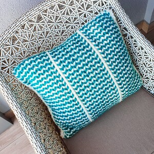 May include: A white wicker chair with a teal and white chevron patterned pillow. The pillow is made of woven fabric and has a soft, plush texture.