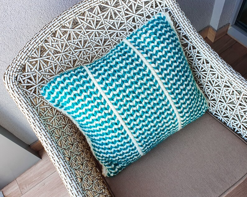 May include: A white wicker chair with a teal and white chevron patterned pillow. The pillow is made of woven fabric and has a soft, plush texture.