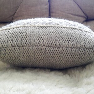 May include: A light grey knitted cushion with a textured surface. The cushion has a rectangular shape with rounded edges and a decorative, lighter-coloured band across the centre. It rests on a white, fluffy surface.