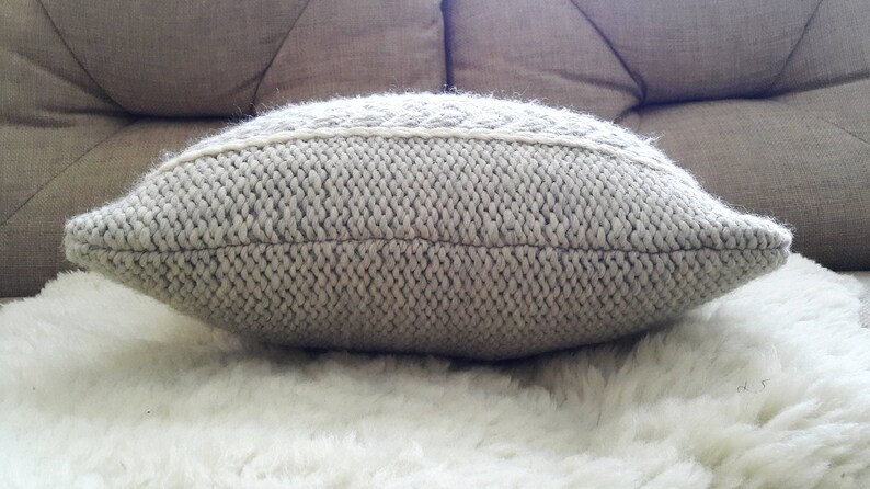 May include: A light grey knitted cushion with a textured surface. The cushion has a rectangular shape with rounded edges and a decorative, lighter-coloured band across the centre. It rests on a white, fluffy surface.