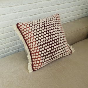 May include: Square decorative pillow with a white and burgundy dotted pattern. The pillow has a fringe trim and is resting on a beige sofa. The pillow's color gradient transitions from white to burgundy.