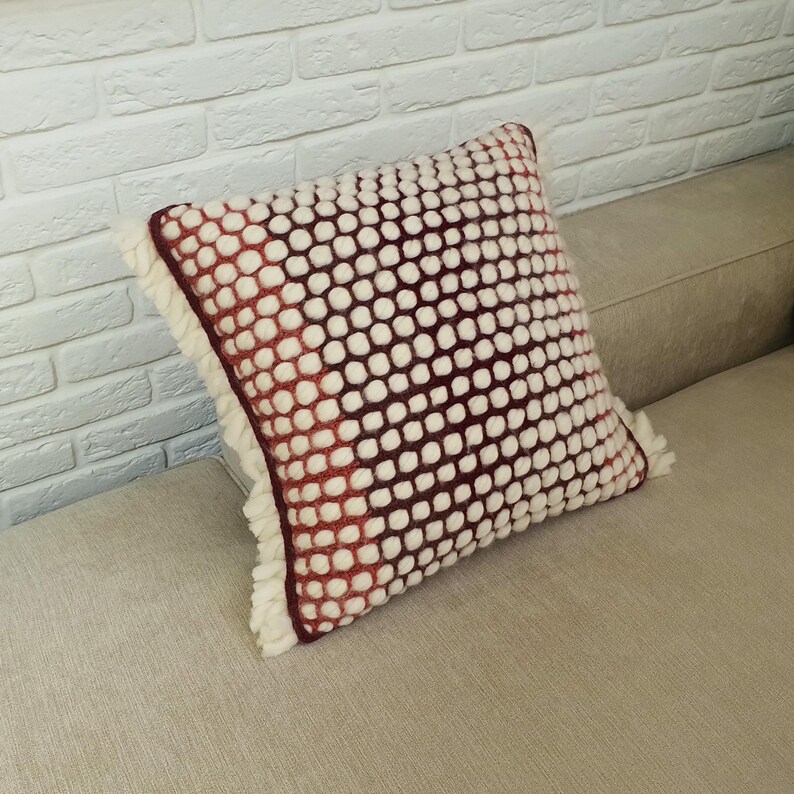 May include: Square decorative pillow with a white and burgundy dotted pattern. The pillow has a fringe trim and is resting on a beige sofa. The pillow's color gradient transitions from white to burgundy.