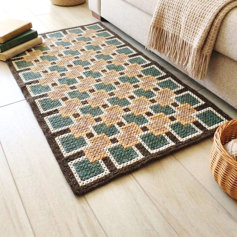 May include: A rectangular, hand-crocheted rug with a geometric pattern. The rug features a repeating design of squares in shades of brown, teal, and beige, outlined in white and framed by a dark brown border. The rug is placed on a light wood floor.