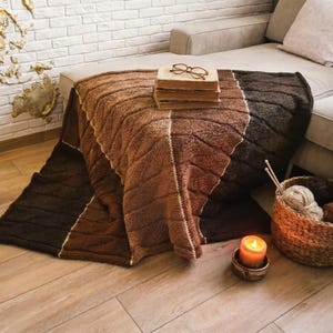 A brown, beige, and dark brown knitted throw blanket with a cable knit pattern. The blanket is folded and placed on a wooden floor.