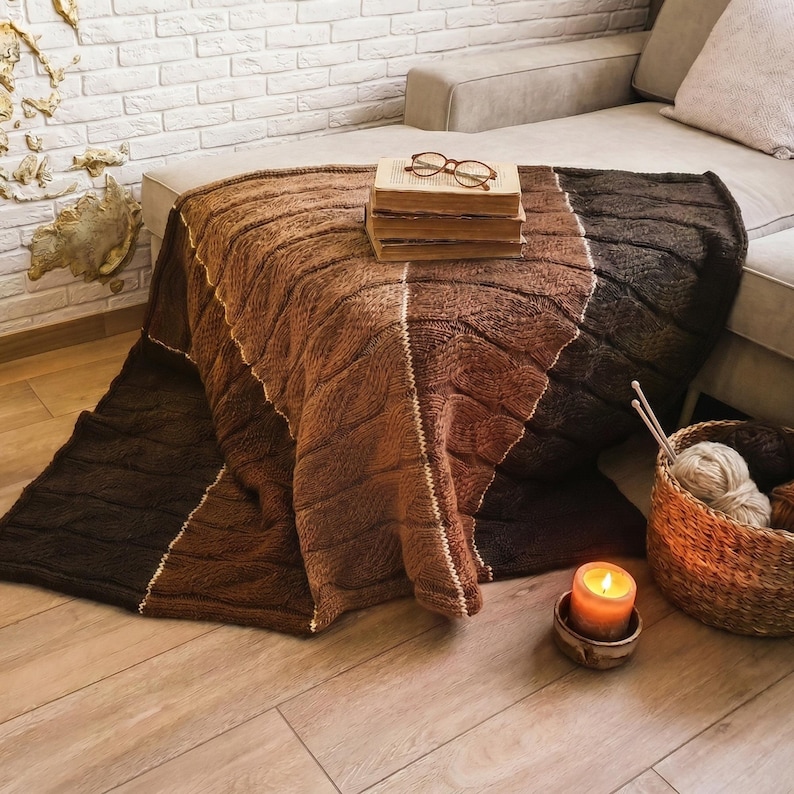 A brown, beige, and dark brown knitted throw blanket with a cable knit pattern. The blanket is folded and placed on a wooden floor.